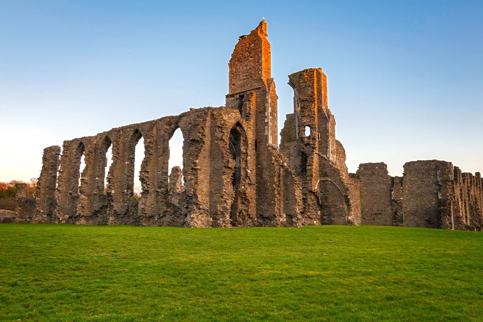 The ruined nave arcade and twin towers of Neath Abbey glowing in evening light, with surviving Cistercian stonework rising above a grass lawn