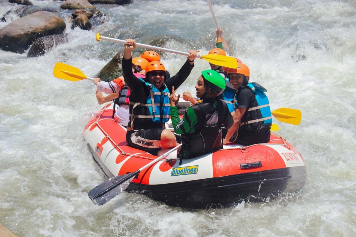 A group of people wearing helmets and life jackets are joyfully white water rafting in Wales, paddling a red inflatable raft through rough, splashing river rapids.