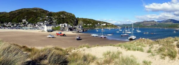 Panoramic view of Barmouth Beach at low tide with grounded boats on the sand under a bright blue sky.
