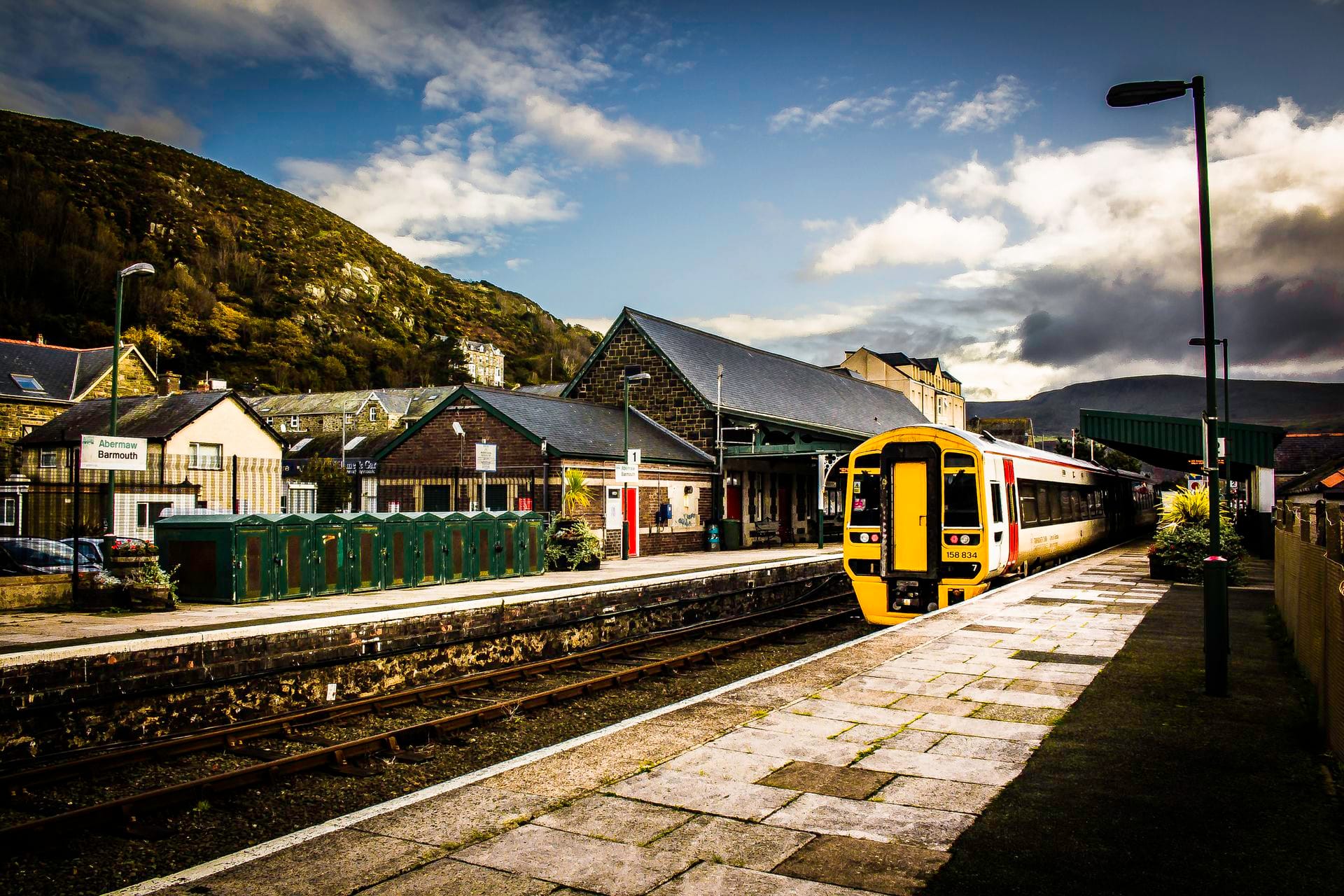 A yellow and white passenger train stopped at the platform of Barmouth railway station. Behind the dark stone station buildings, a steep, green hillside rises against a partly cloudy sky. A sign on the opposite platform reads