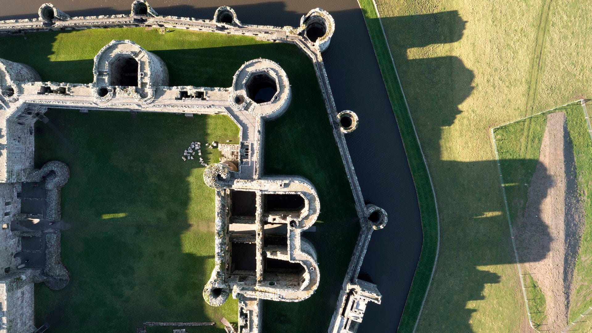 An aerial view of Beaumaris Castle showing its concentric design, with a ring of corner towers and a curtain wall enclosing the grassed inner ward, and a moat running along one side.