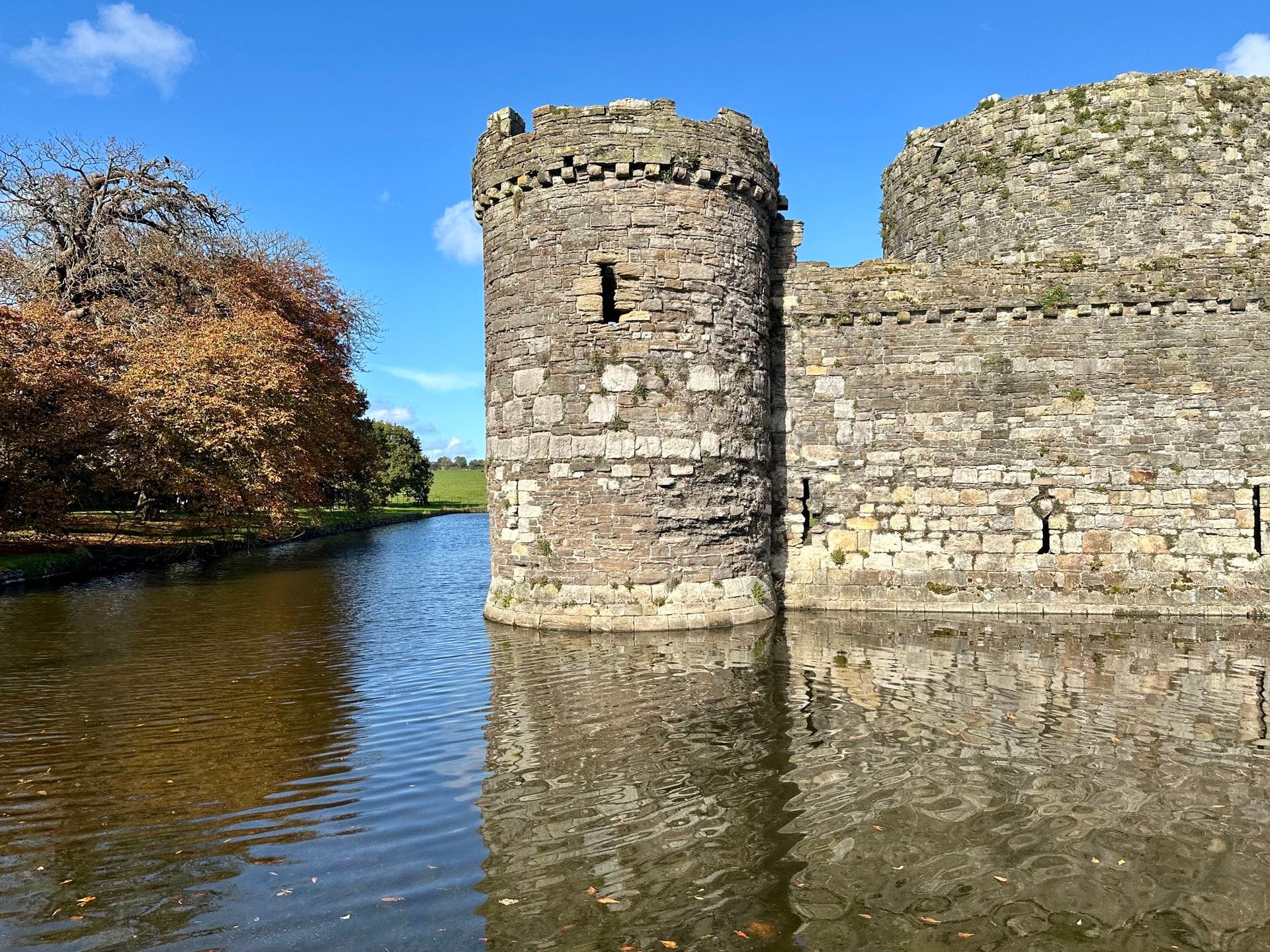Beaumaris Castle's curtain wall and two round towers seen across the water-filled moat, with green fields visible beyond the walls.