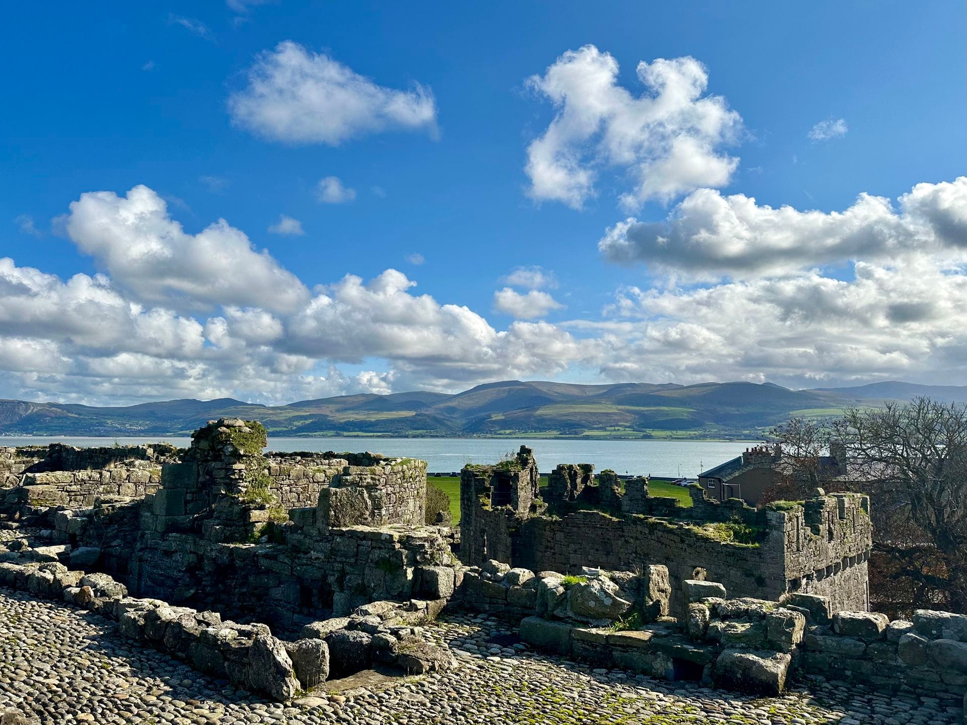The low ruined walls of Beaumaris Castle's outer defences, with a wide stretch of water and the mountains of the Welsh mainland visible in the distance under a partly cloudy sky.
