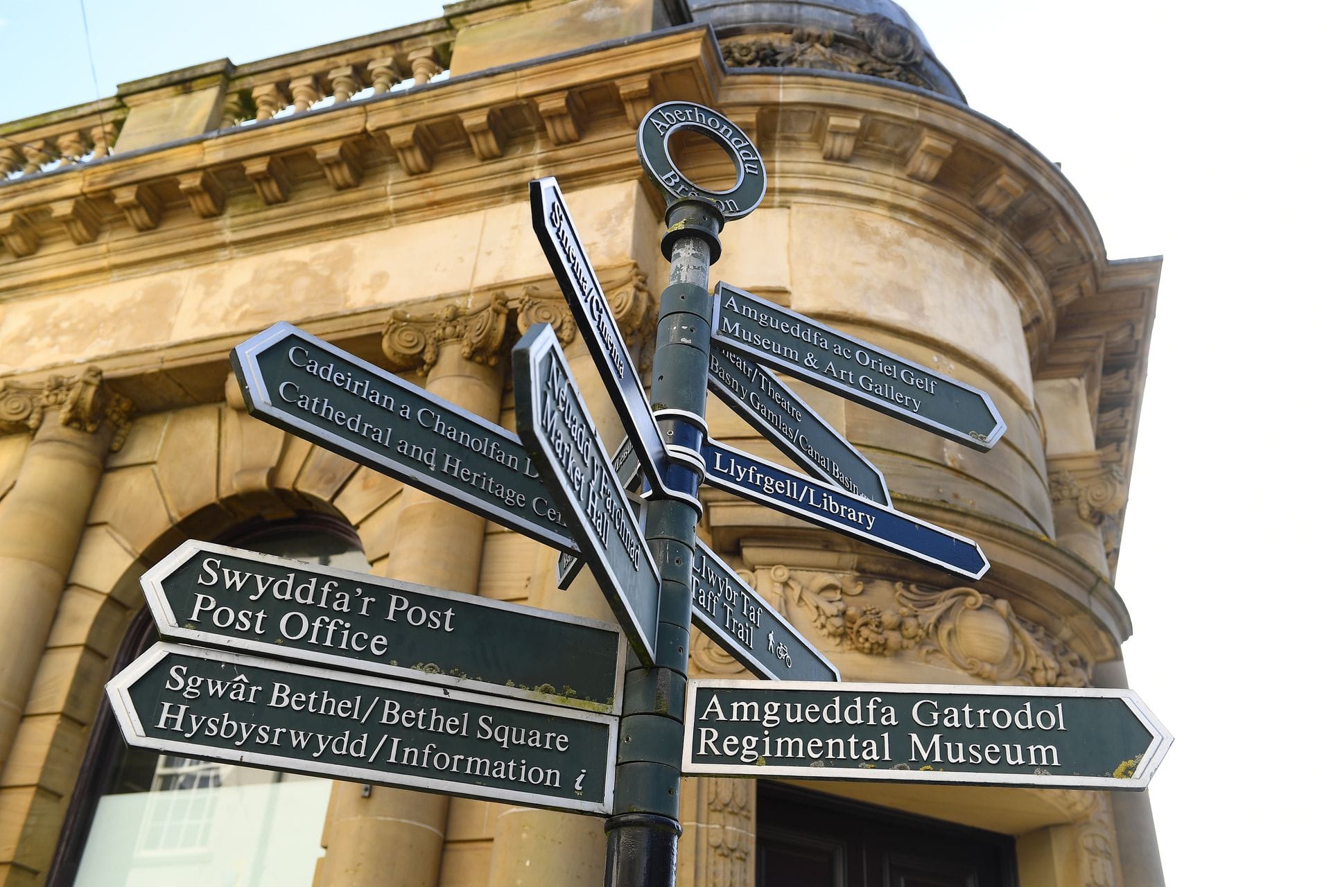 A black metal directional fingerpost with several signs pointing in different directions. The signs feature white text displaying town locations in both Welsh and English, including "Swyddfa'r Post / Post Office" and "Amgueddfa Gatrodol / Regimental Museum". The signpost stands in front of a curved, classical stone building with ornate architectural details.