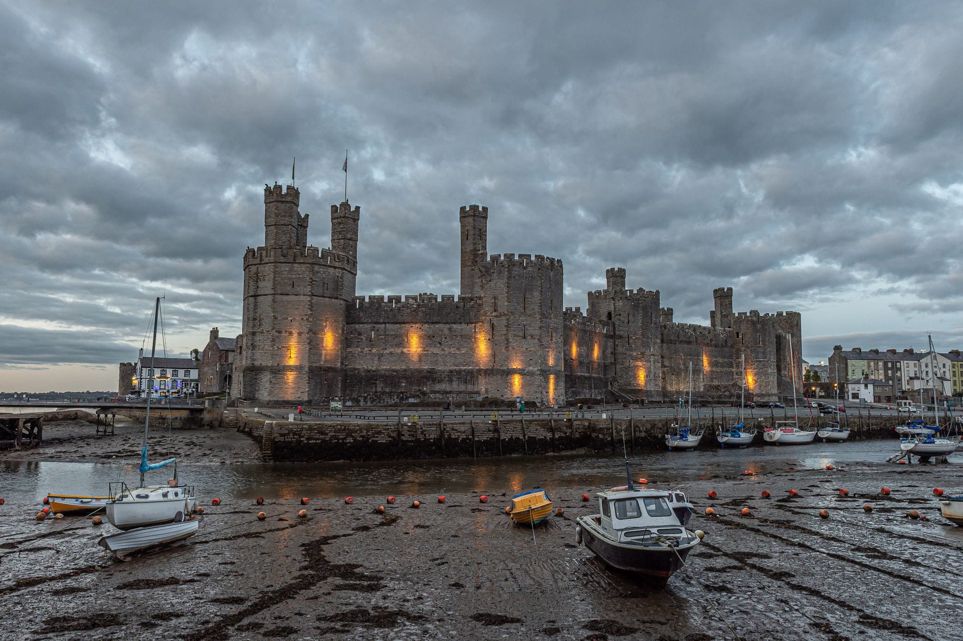 Caernarfon Castle illuminated in warm light at dusk, viewed from the harbour with small boats grounded on the mud at low tide and a dramatic cloudy sky above.