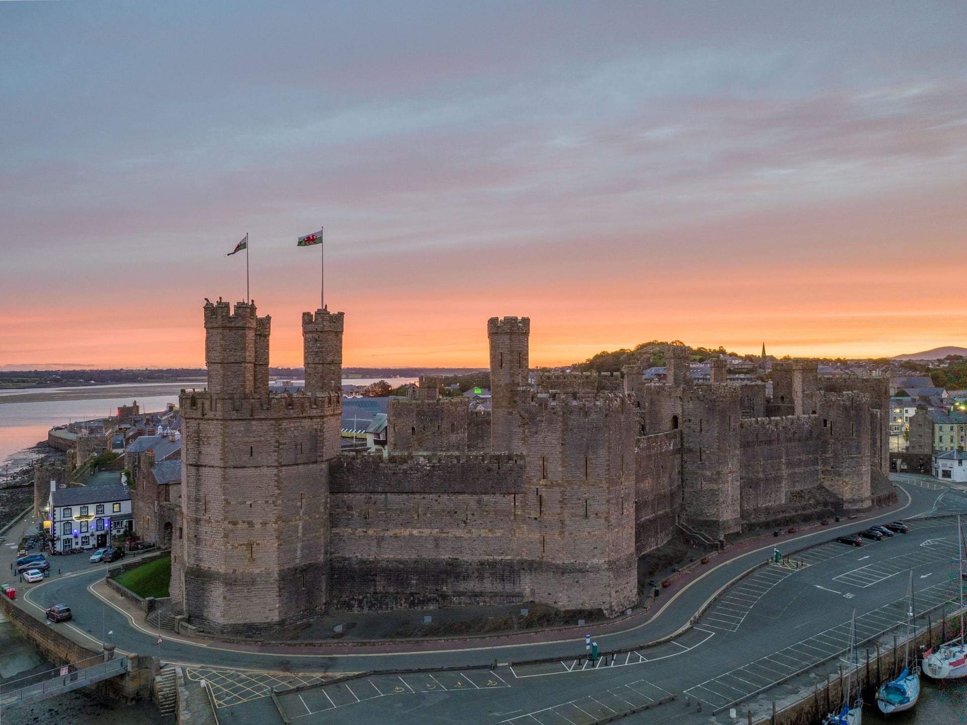 Caernarfon Castle's turreted towers silhouetted against a vivid orange sunset sky, with two Welsh flags flying from the battlements and the town and estuary visible below