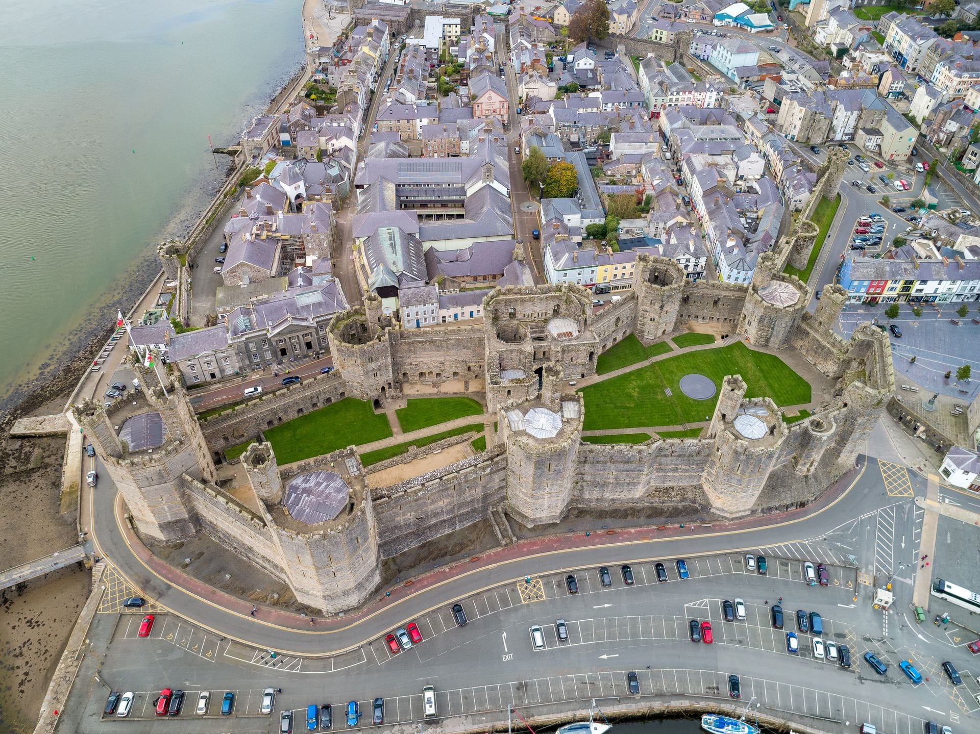 An aerial view of Caernarfon Castle's full curtain wall and polygonal towers alongside the Menai Strait, with the medieval walled town, colourful shopfronts and car parks surrounding the fortress.