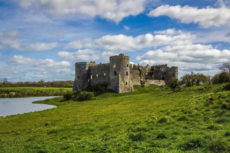 Carew Castle with round towers stands on a grassy hill by a small lake, perfect for short breaks in North Wales. Surrounded by green fields and trees under a partly cloudy blue sky, it offers a peaceful escape.
