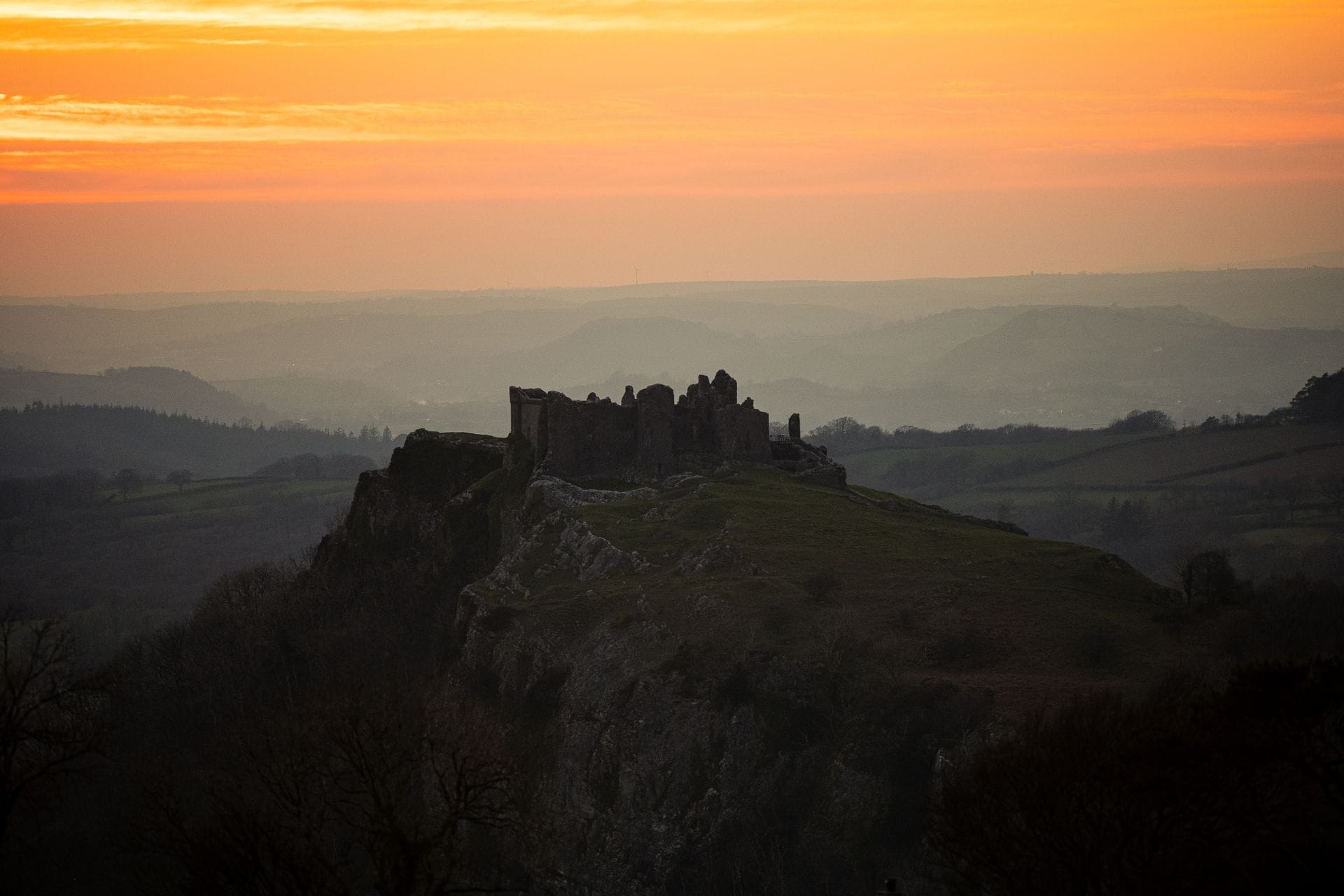 Silhouette of Carreg Cennen Castle ruins perched on a rocky crag against a bright orange sunset over misty rolling hills