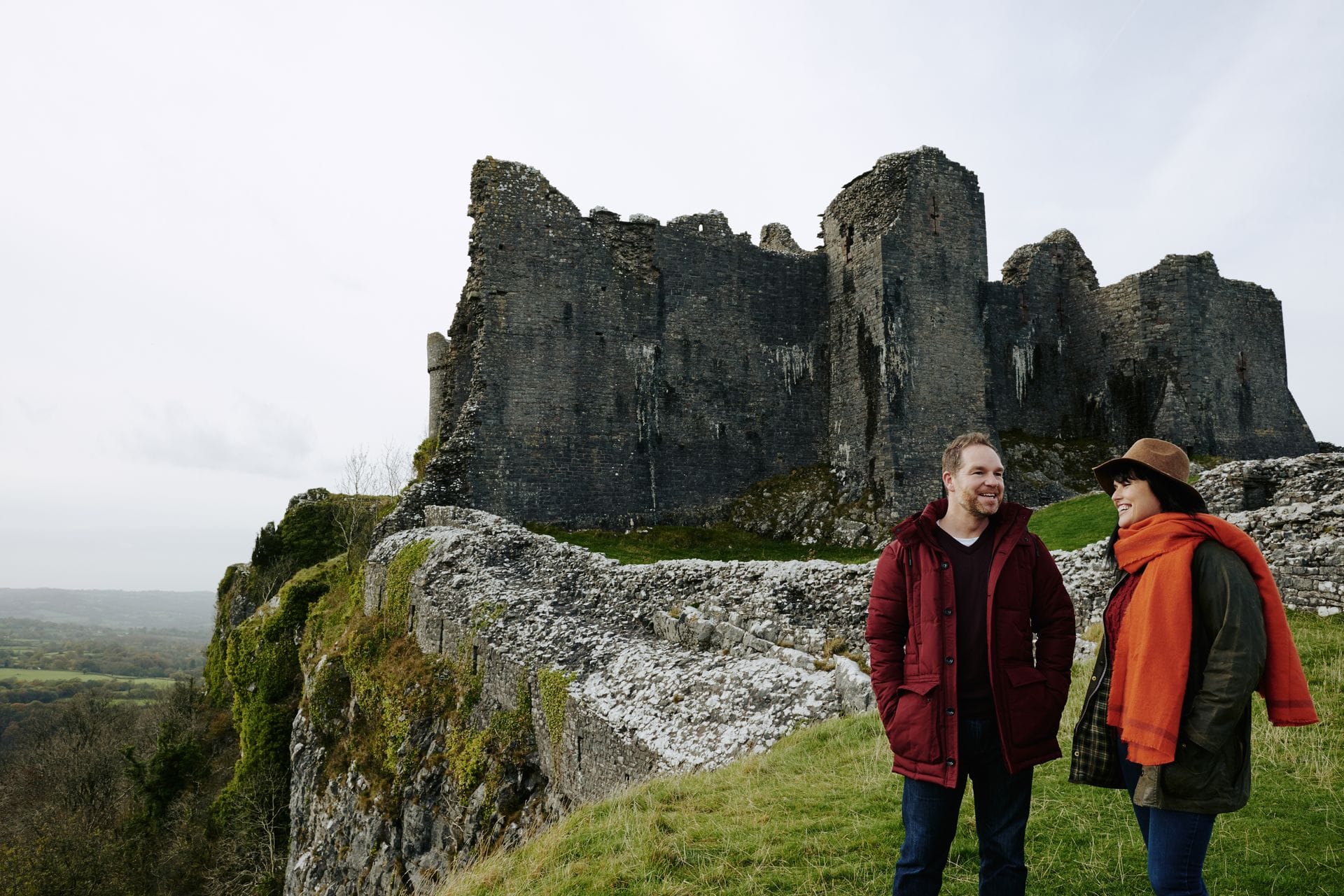 A man and a woman standing on the grassy edge of Carreg Cennen Castle's cliff, with the towering stone ruins directly behind them and a vast green valley below.