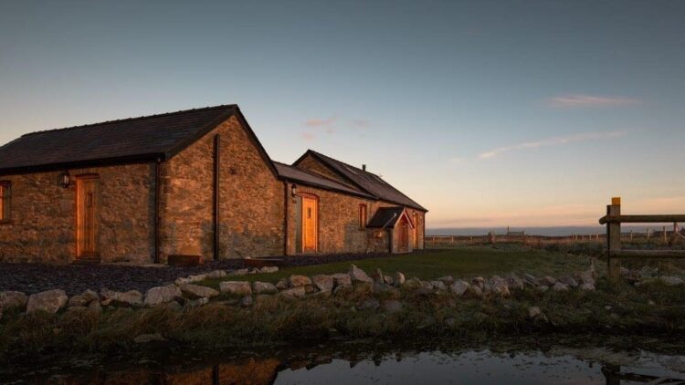 A rustic stone cottage with a slate roof is illuminated by warm sunlight at sunset, surrounded by grass, rocks, and a small pond—one of the charming dog-friendly holiday cottages in Anglesey with countryside views and a peaceful pale sky beyond.