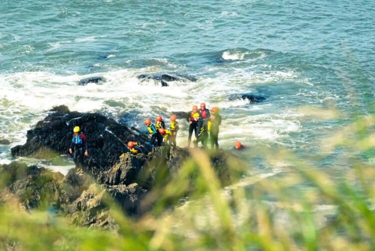 A group of people wearing helmets and wetsuits stand and sit on rocks by the sea, coasteering Wales style, with waves crashing around them. Some appear to be helping others. Green grass is visible in the foreground.