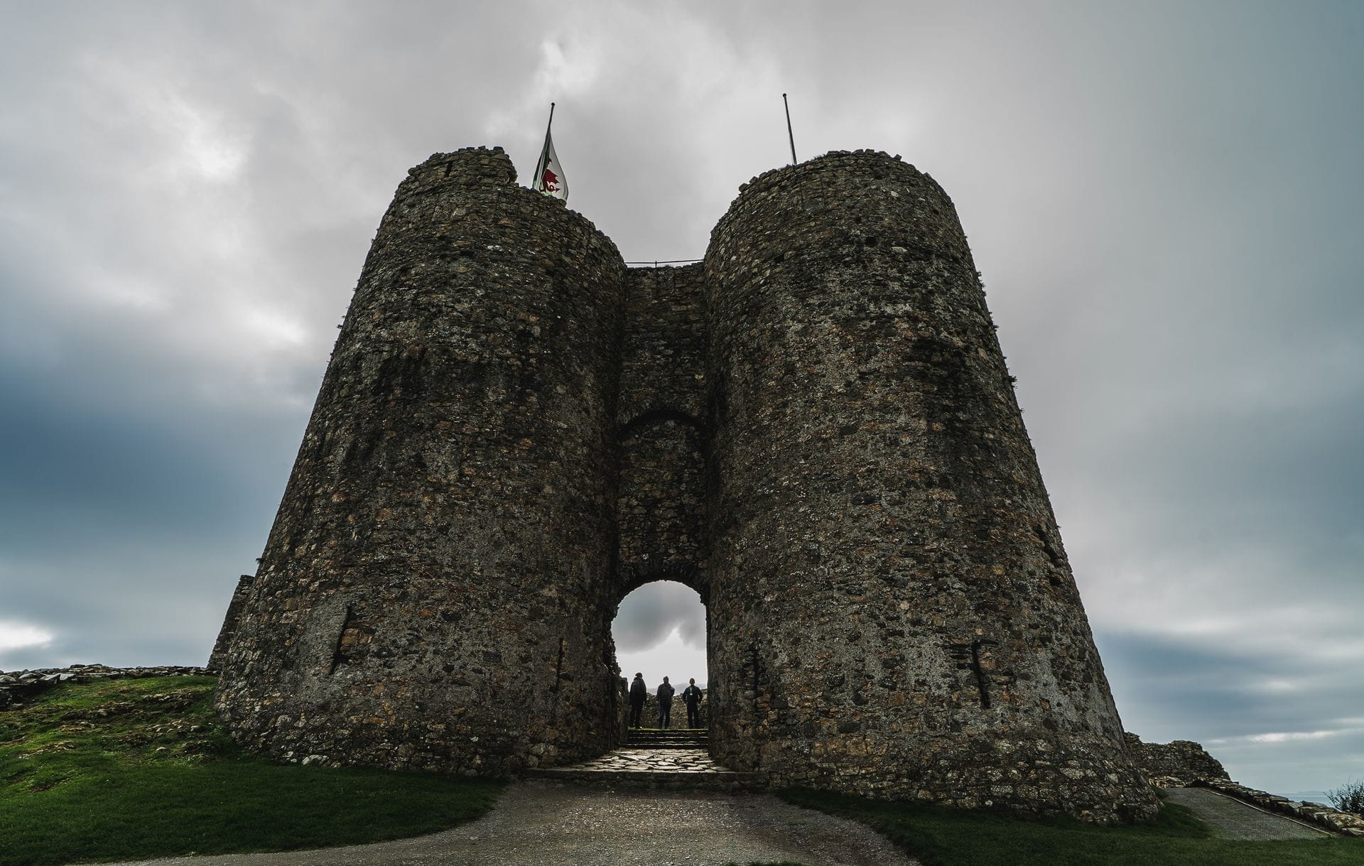 A low-angle view looking up at Criccieth Castle's twin D-shaped gatehouse towers, with visitors walking through the stone archway between them and a Welsh flag flying above.