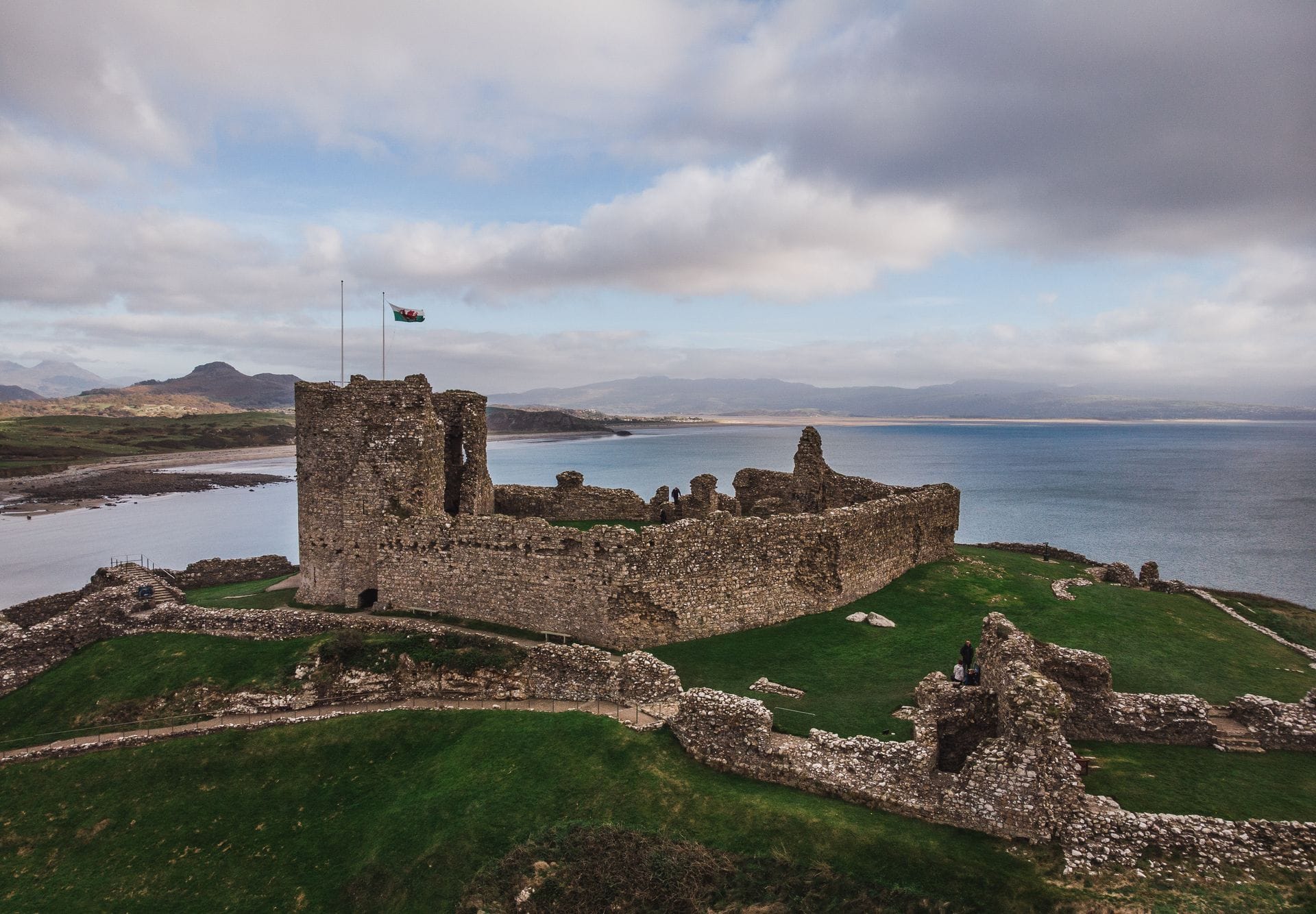The silhouette of Criccieth Castle atop its headland at sunset, with layered mountains and Tremadog Bay fading into golden haze behind it.