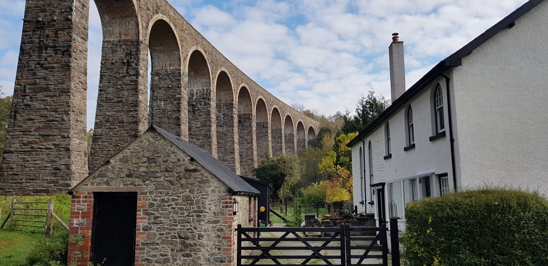 A tall, curved stone railway viaduct with multiple arches stretching across a valley under a blue sky, with a white house and farm gate in the foreground