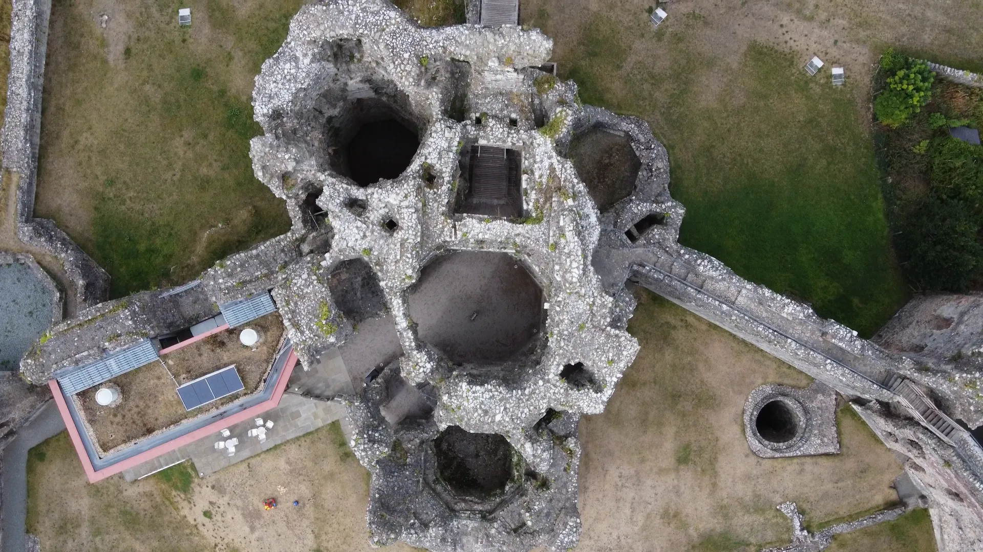 An aerial view directly above Denbigh Castle's ruined triple-towered gatehouse, showing the polygonal shapes of the towers clustered together, with a modern visitor centre building and green lawns visible alongside.