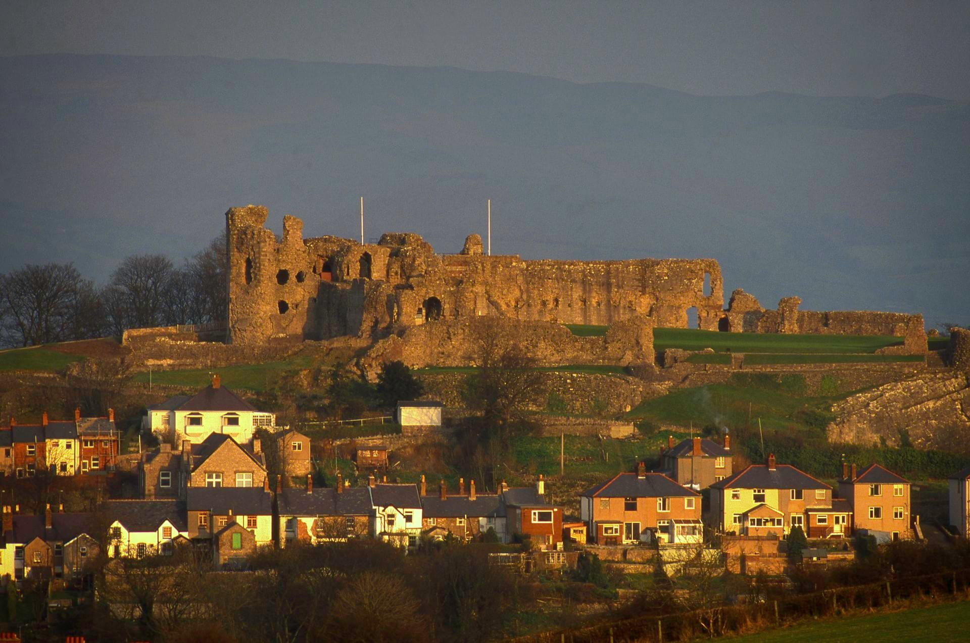 Denbigh Castle's ruined walls catching golden evening light on a hilltop above the town of Denbigh, with rows of houses in the foreground and hills fading into haze behind.