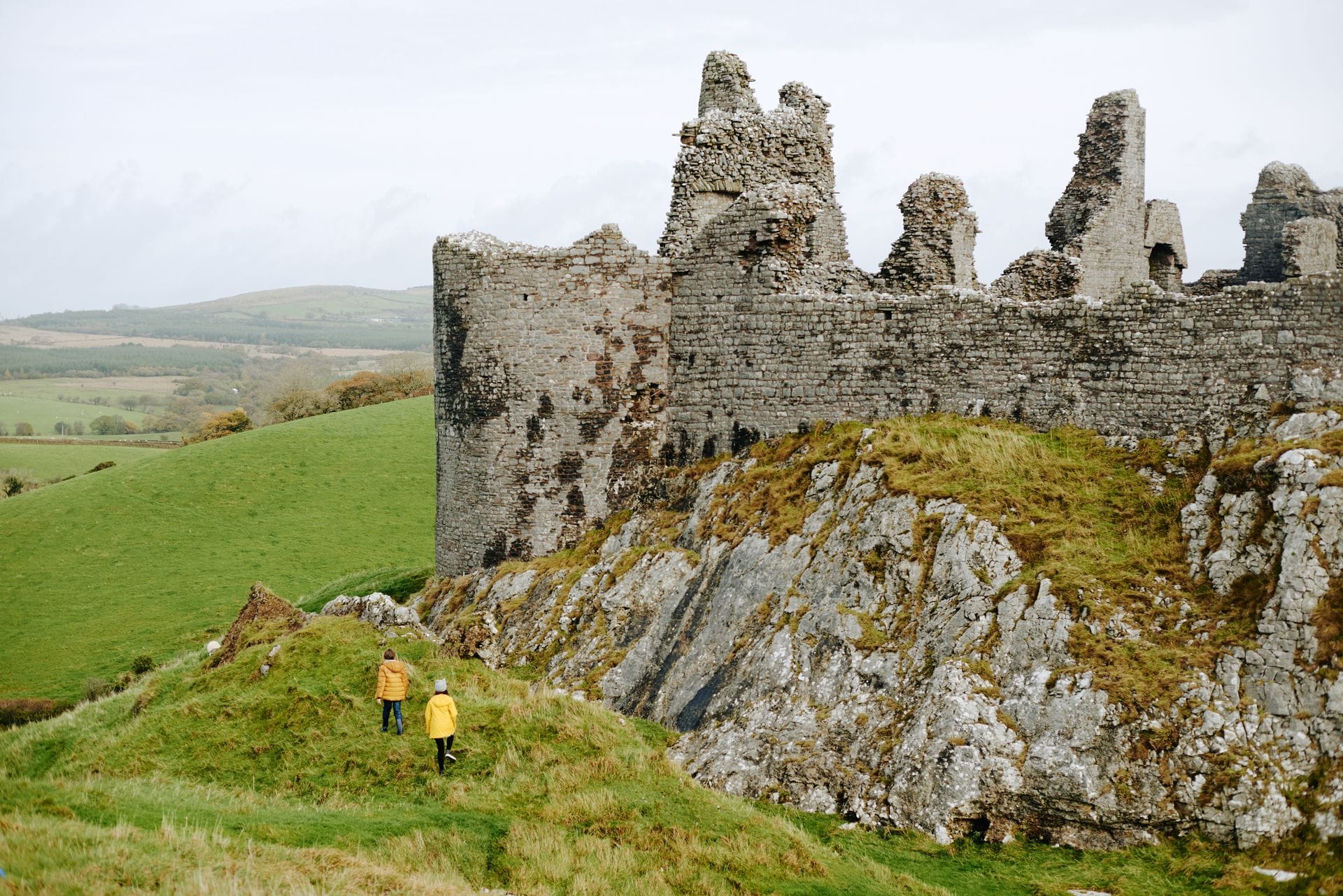 Two children in bright coats walk up a grassy slope toward the towering stone ruins of Carreg Cennen Castle, which sits on a jagged limestone foundation