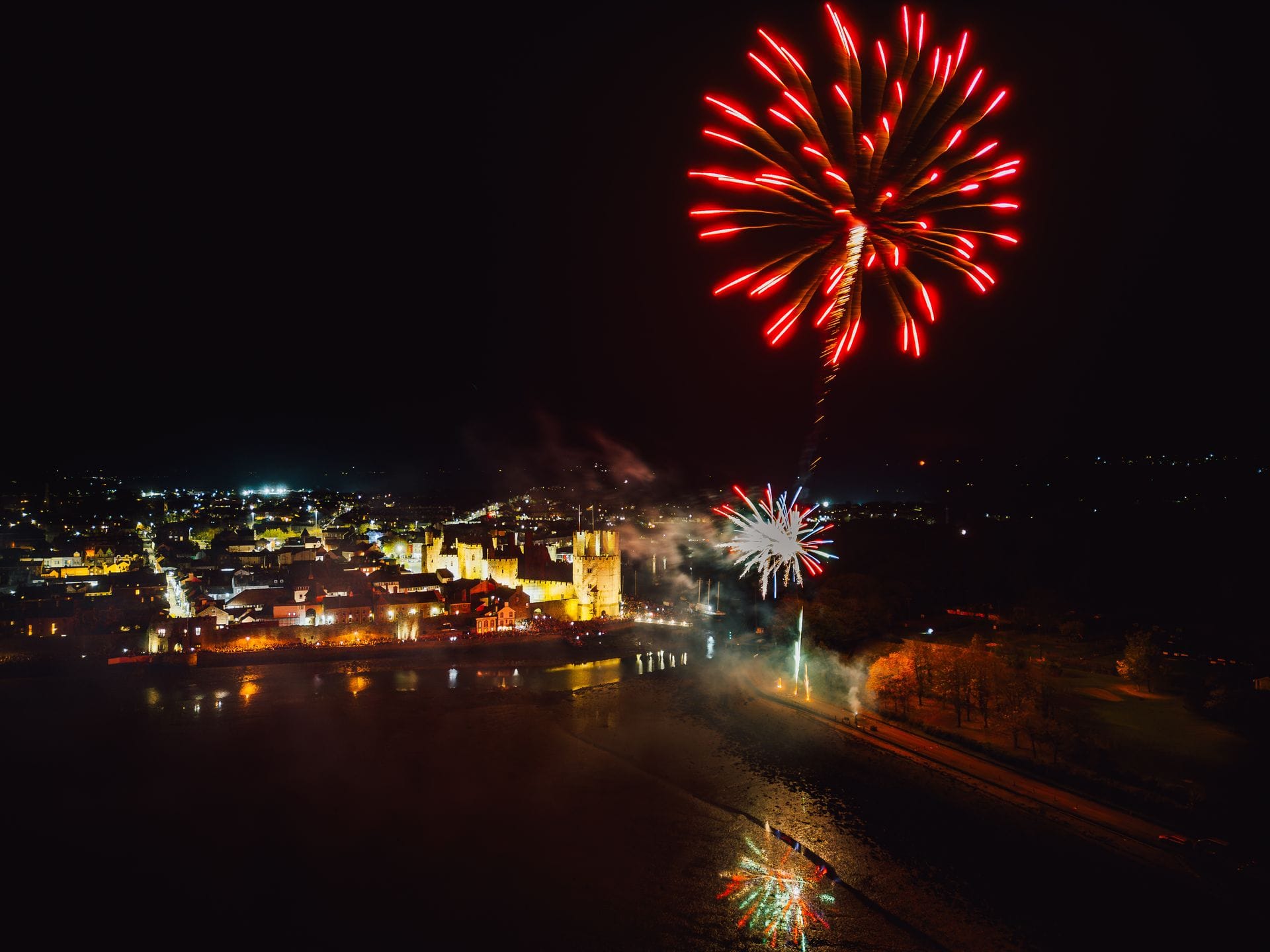 A large red firework explodes high in the night sky over the warmly illuminated stone walls and polygonal towers of Caernarfon Castle. The fortress and surrounding town sit beside a dark body of water that reflects the town lights and smaller fireworks bursting closer to the ground.
