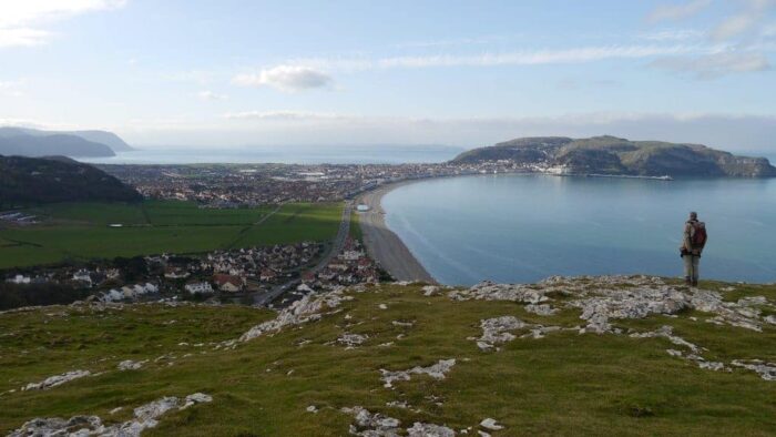 A person stands on a grassy hill overlooking the coastal town and the sweeping bay beneath Great Orme, with distant hills under a partly cloudy sky. The sea glistens in the sunlight, and houses dot the shore.