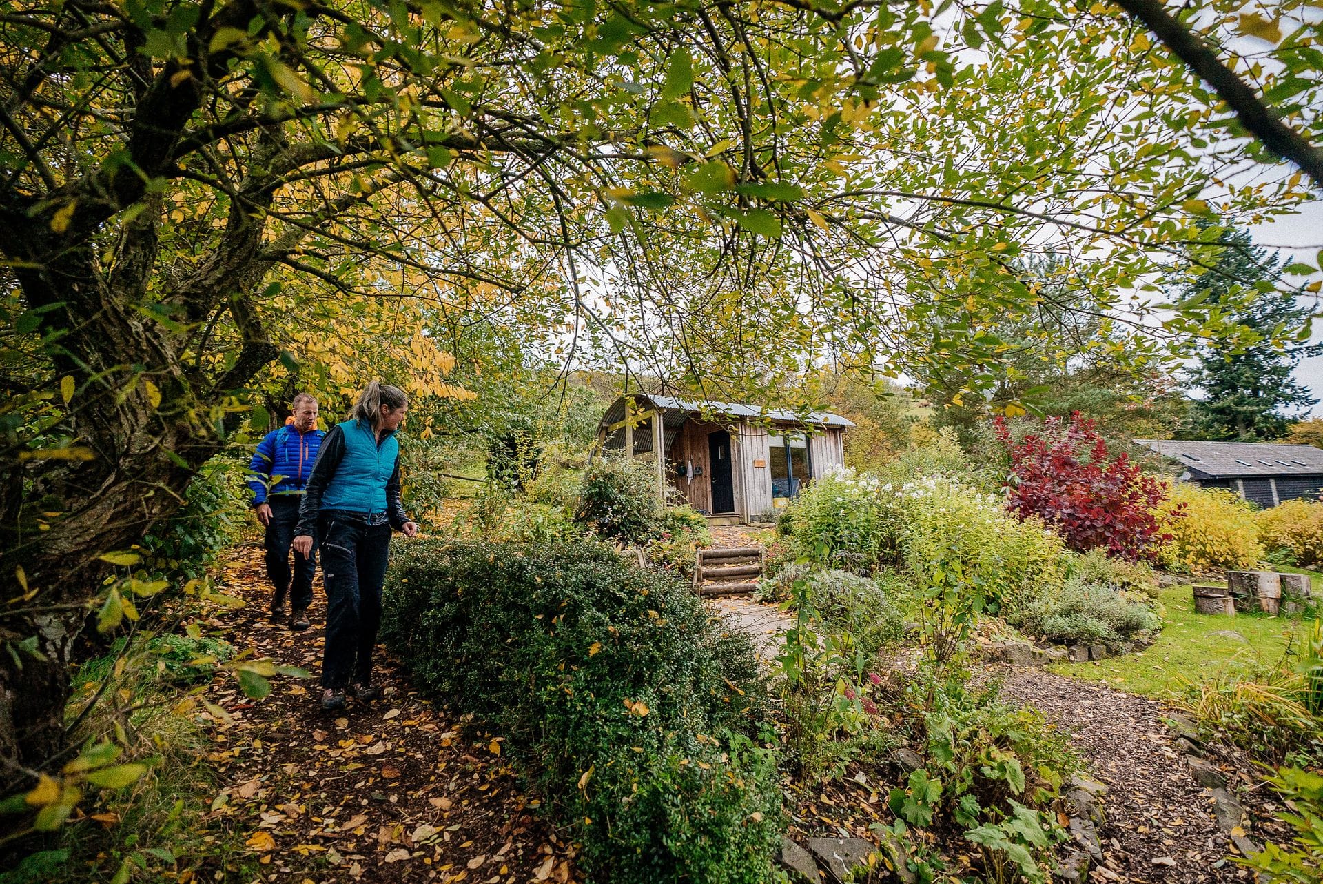 Two hikers in outdoor gear walking along a gravel path through a lush, mature garden towards a wooden guesthouse on a Welsh walking holiday.