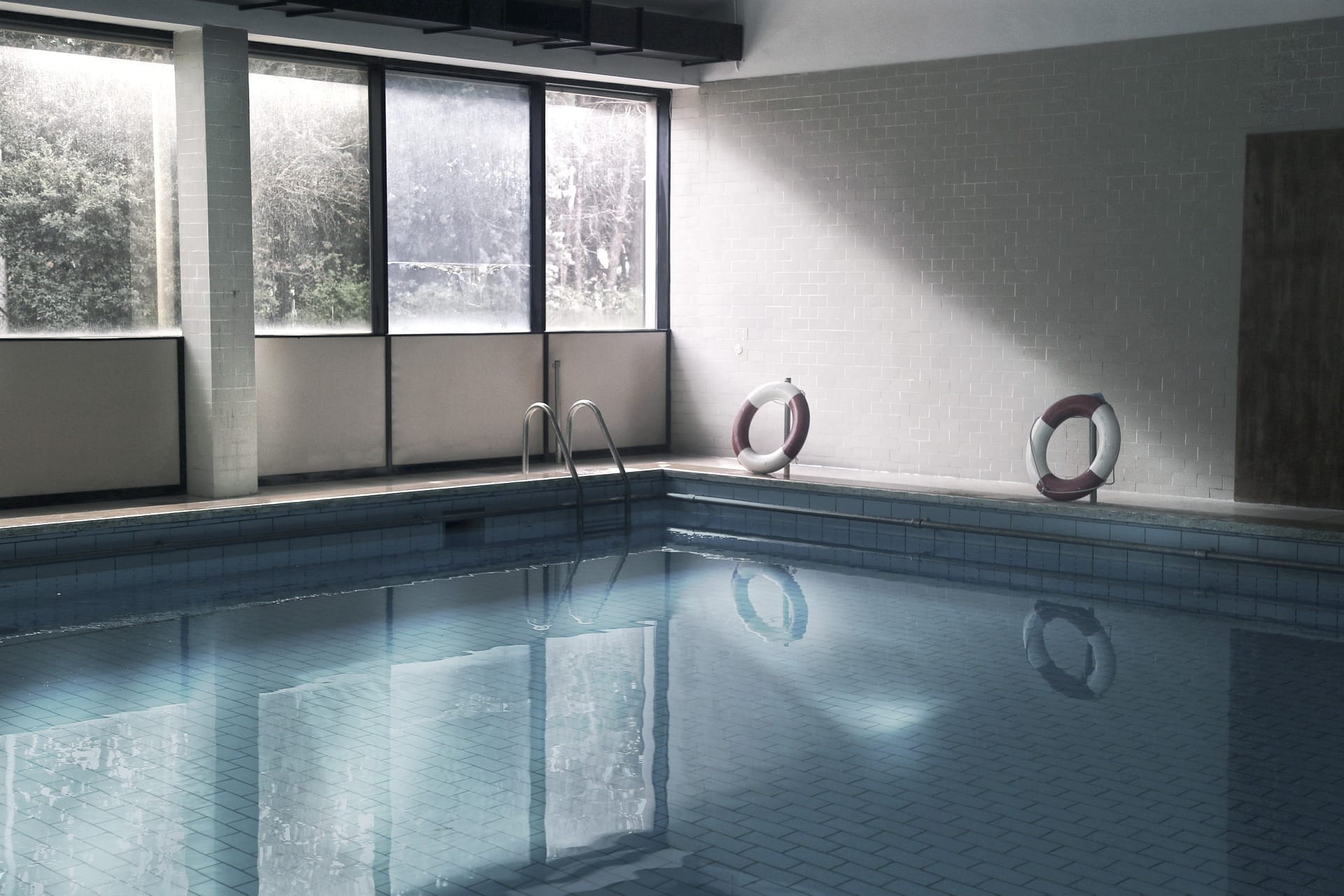 An atmospheric indoor swimming pool with pale blue tiled water, tall industrial-style windows on the left overlooking bare winter trees, white-brick walls, two red-and-white life ring buoys placed on the pool deck, and a chrome ladder descending into the still water — an example of an indoor cottage or country-house pool giving guaranteed weatherproof swimming through the Welsh winter and shoulder seasons.