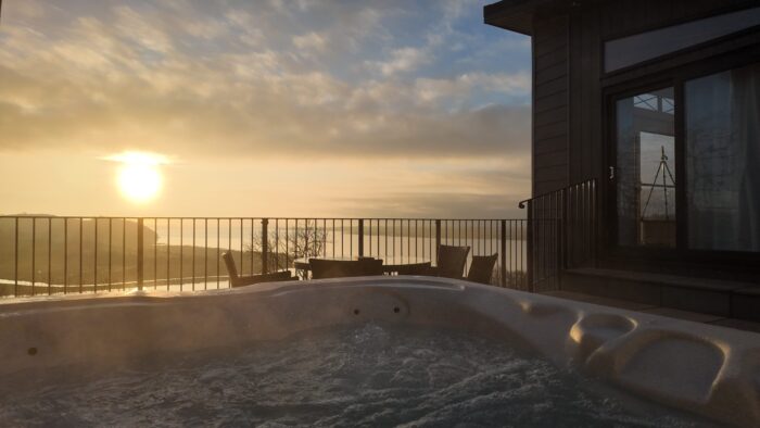 A hot tub with bubbling water sits on a patio overlooking a scenic sunset—one of the charming cottages with hot tubs in Wales. A table and chairs by the balustrade invite you to relax as the sun casts a warm glow over the landscape and house.