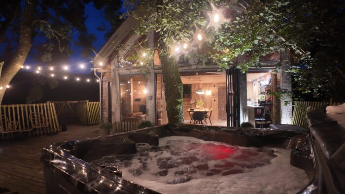 A cosy wooden cabin at night is warmly lit inside and outside with fairy lights. In the foreground, a bubbling hot tub sits on a wooden decking—perfect for a relaxing hot tub holiday in Wales—surrounded by trees and rustic outdoor furniture.