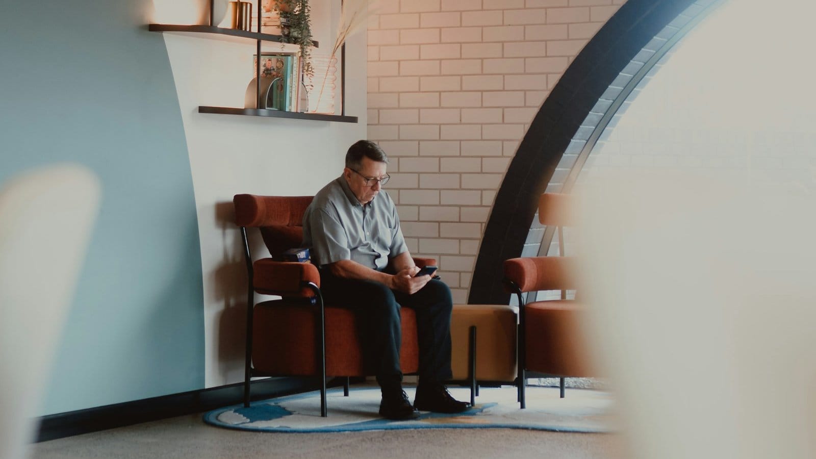A man in glasses sitting in a rust-coloured armchair in a modern lounge, looking at his phone, with a white brick wall and a large arched window behind him.