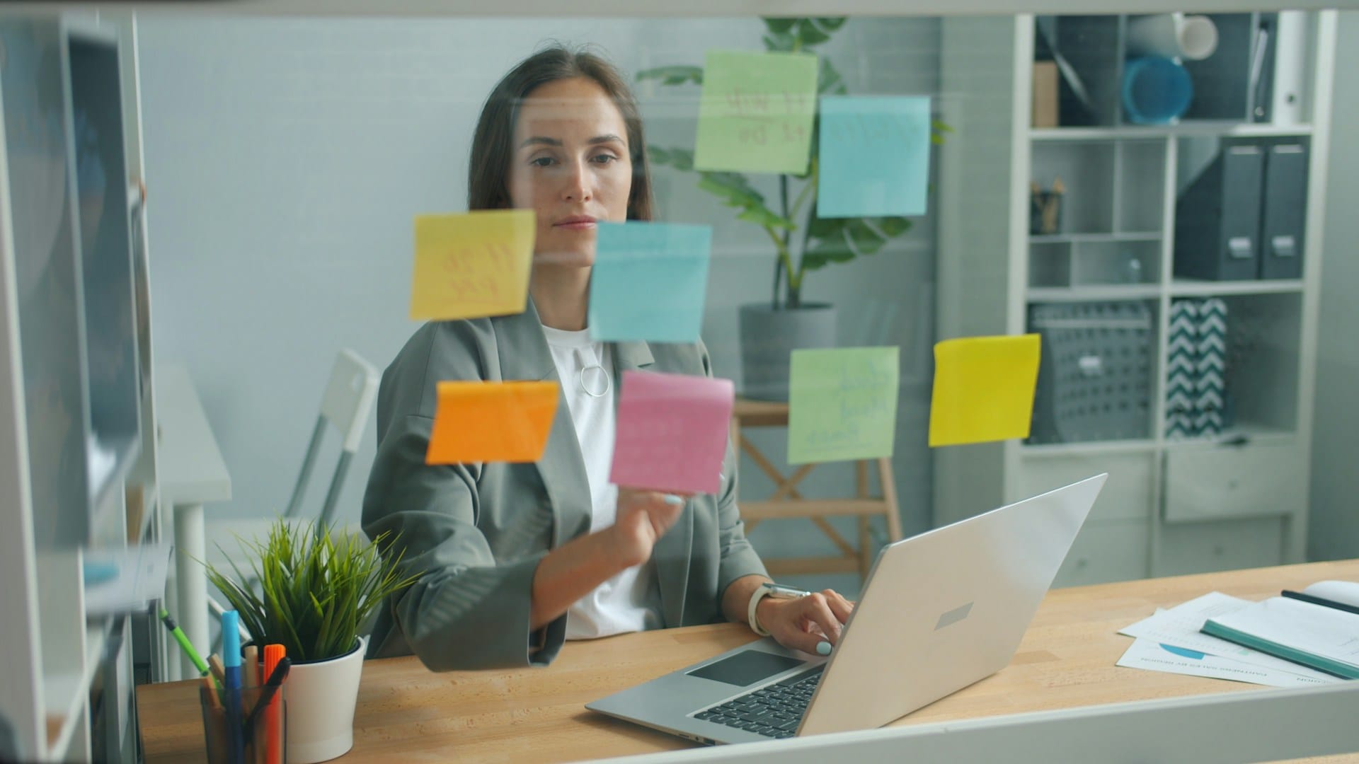 A woman in a grey blazer at a desk with a laptop, placing coloured sticky notes on a glass board