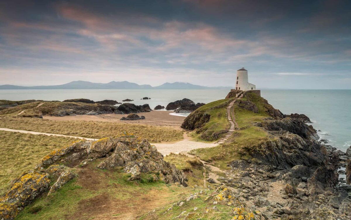 A winding sandy path leading to the historic white Tŵr Mawr lighthouse, which sits perfectly perched on a rocky, grass-covered headland on Llanddwyn Island. The calm sea and distant mountains of Eryri form a dramatic backdrop under a cloudy sky.