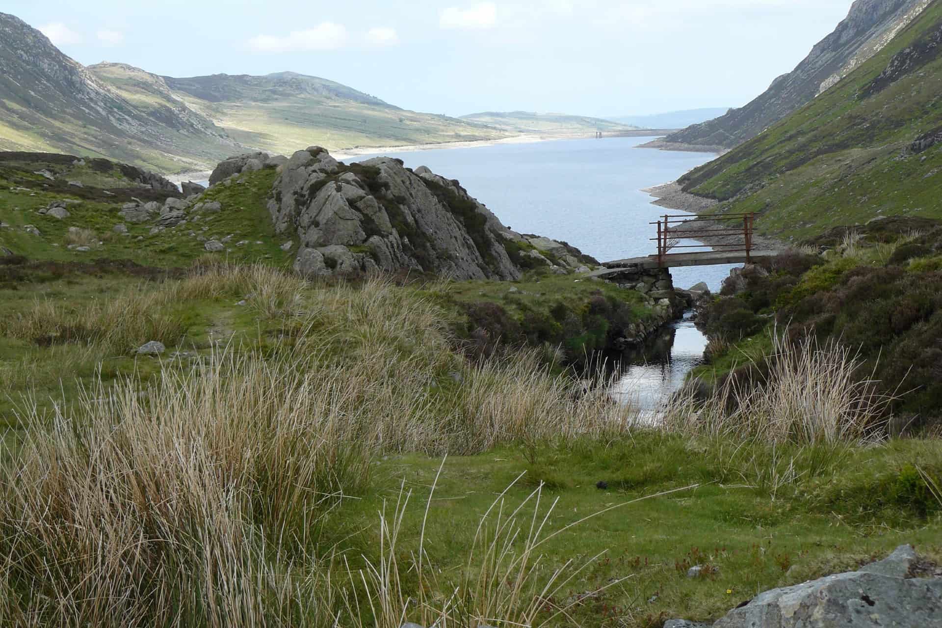A scenic wilderness view in the South Wales Valleys featuring a calm blue lake nestled between rolling, grassy mountains, with a small wooden footbridge crossing a narrow stream in the rocky, grass-covered foreground.