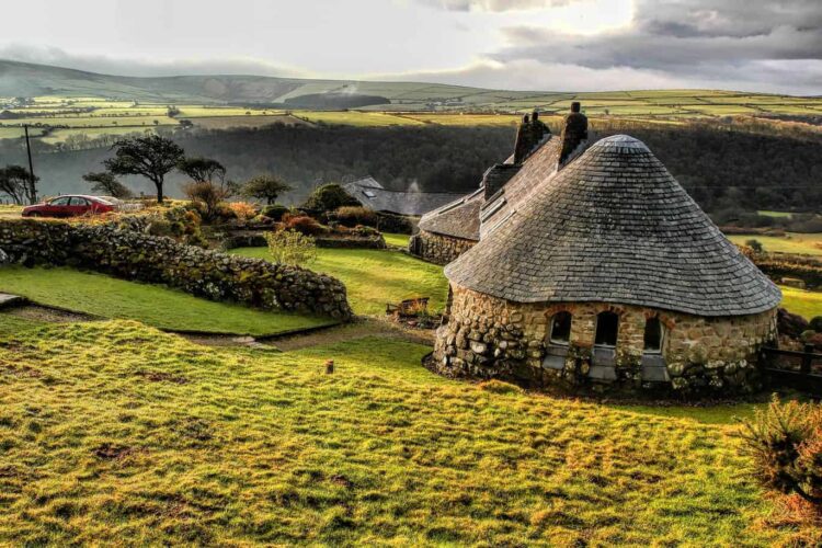 A stone cottage with a conical slate roof sits on a grassy hillside, surrounded by stone walls and gardens—perfect for working farm holidays in Wales, with rolling hills and fields visible under a cloudy sky.