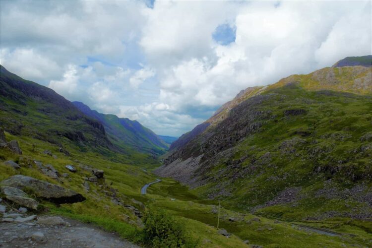 A winding road runs through a lush green valley between rugged, rocky mountains under a partly cloudy sky, capturing the wild beauty of Welsh landscapes and the unspoilt charm of nature in Wales.