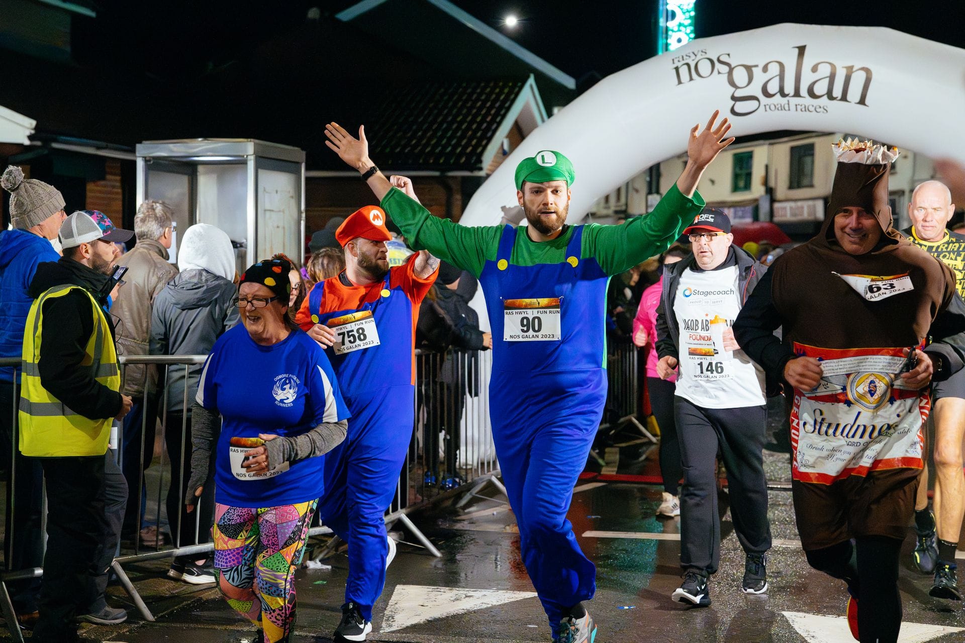 A group of runners, including several in fancy dress costumes like Mario, Luigi, and a pint glass, run along a wet street at night under a white inflatable arch reading