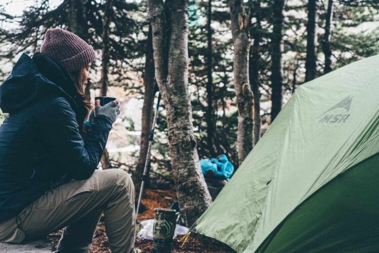 A person wearing a beanie and jacket sits beside a green tent in a forest, holding a mug. Camping gear is scattered nearby among the trees, capturing the cosy spirit of camping in Wales, surrounded by natural beauty.