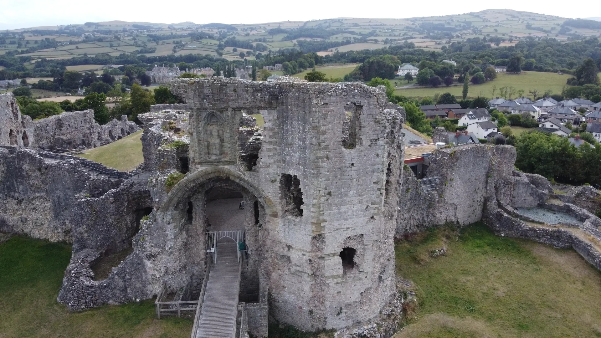 An aerial view of Denbigh Castle's ruined gatehouse with a wooden walkway crossing into the entrance archway, the town of Denbigh and the rolling countryside of the Vale of Clwyd stretching into the distance beyond.