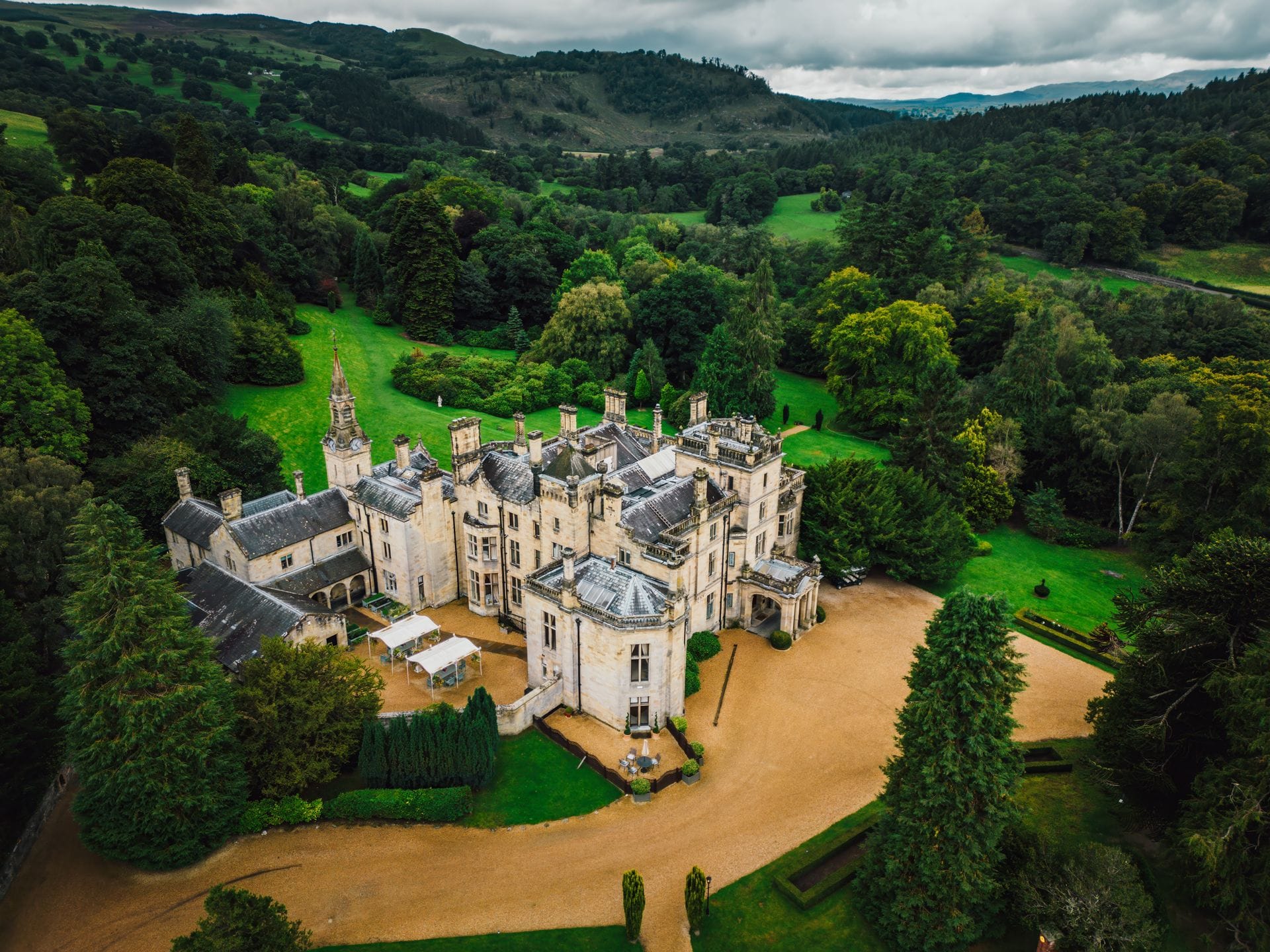 An aerial view of Palé Hall, a grand Victorian country house hotel built from light stone with multiple turrets and chimneys. The estate is surrounded by manicured green lawns, tall trees, and rolling forested hills under a cloudy sky.