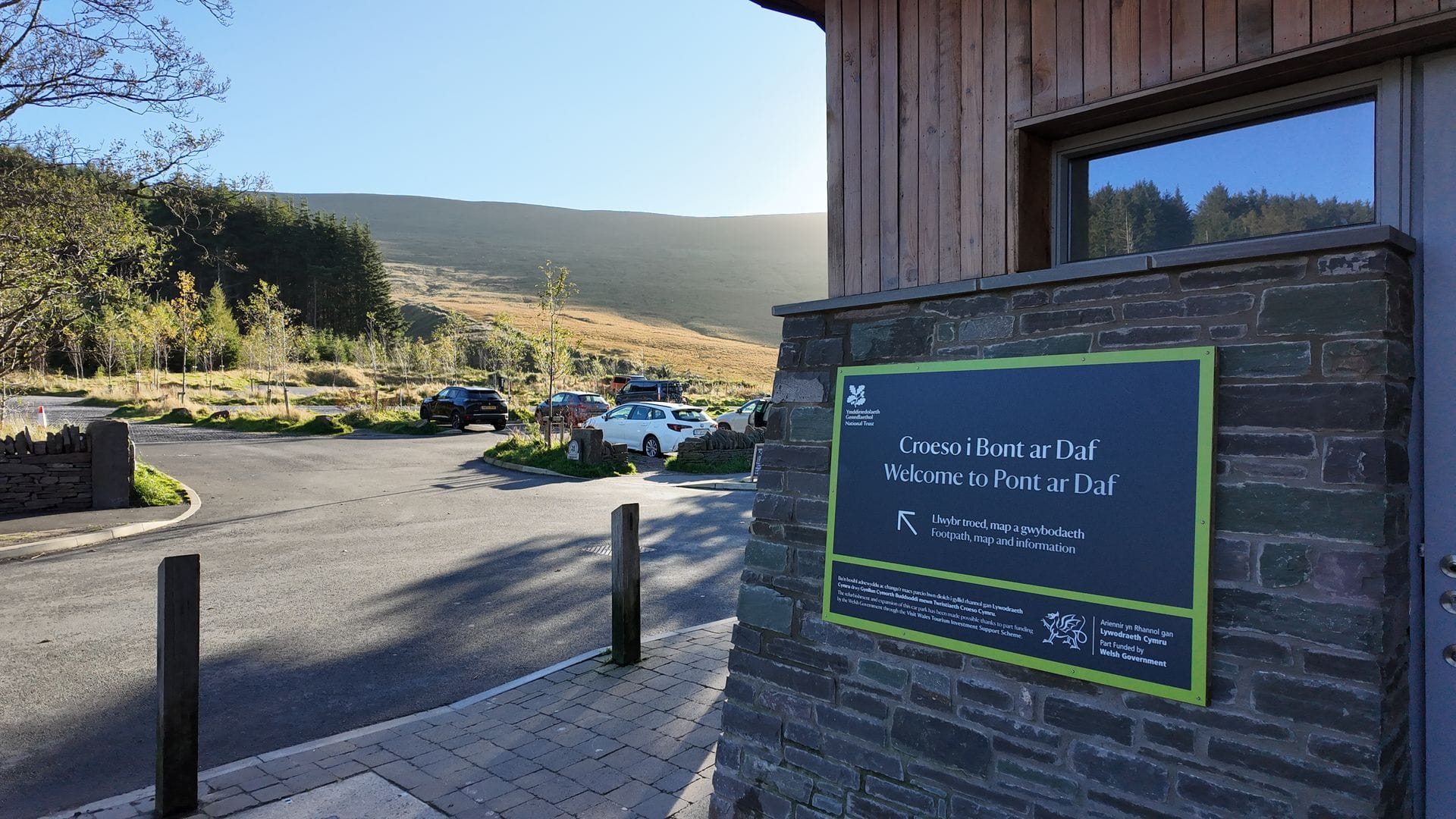Cars parked in the main Pen y Fan visitor car park with a sweeping view of the rolling hills and mountain peaks of the Brecon Beacons in the background.