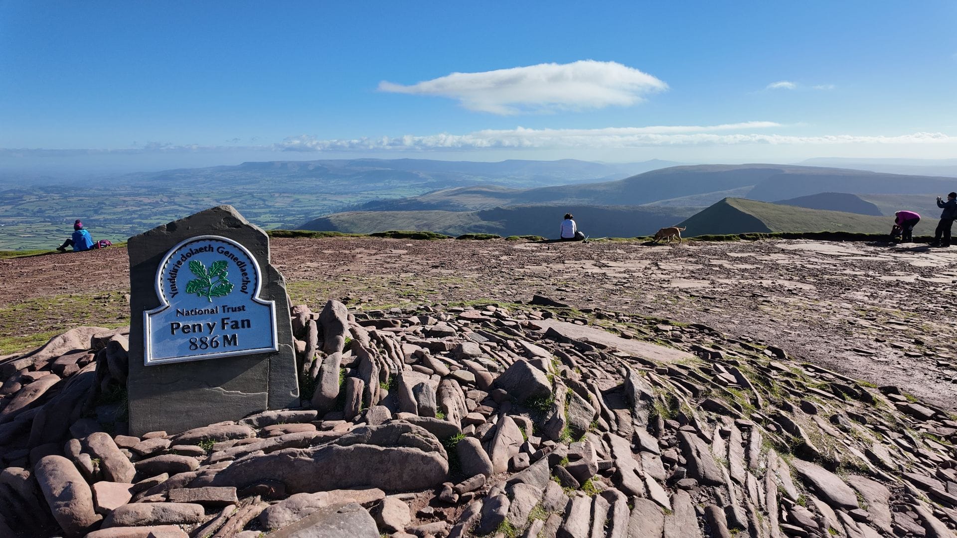 Hikers standing next to the iconic National Trust Pen y Fan summit marker stone surrounded by winter snow.