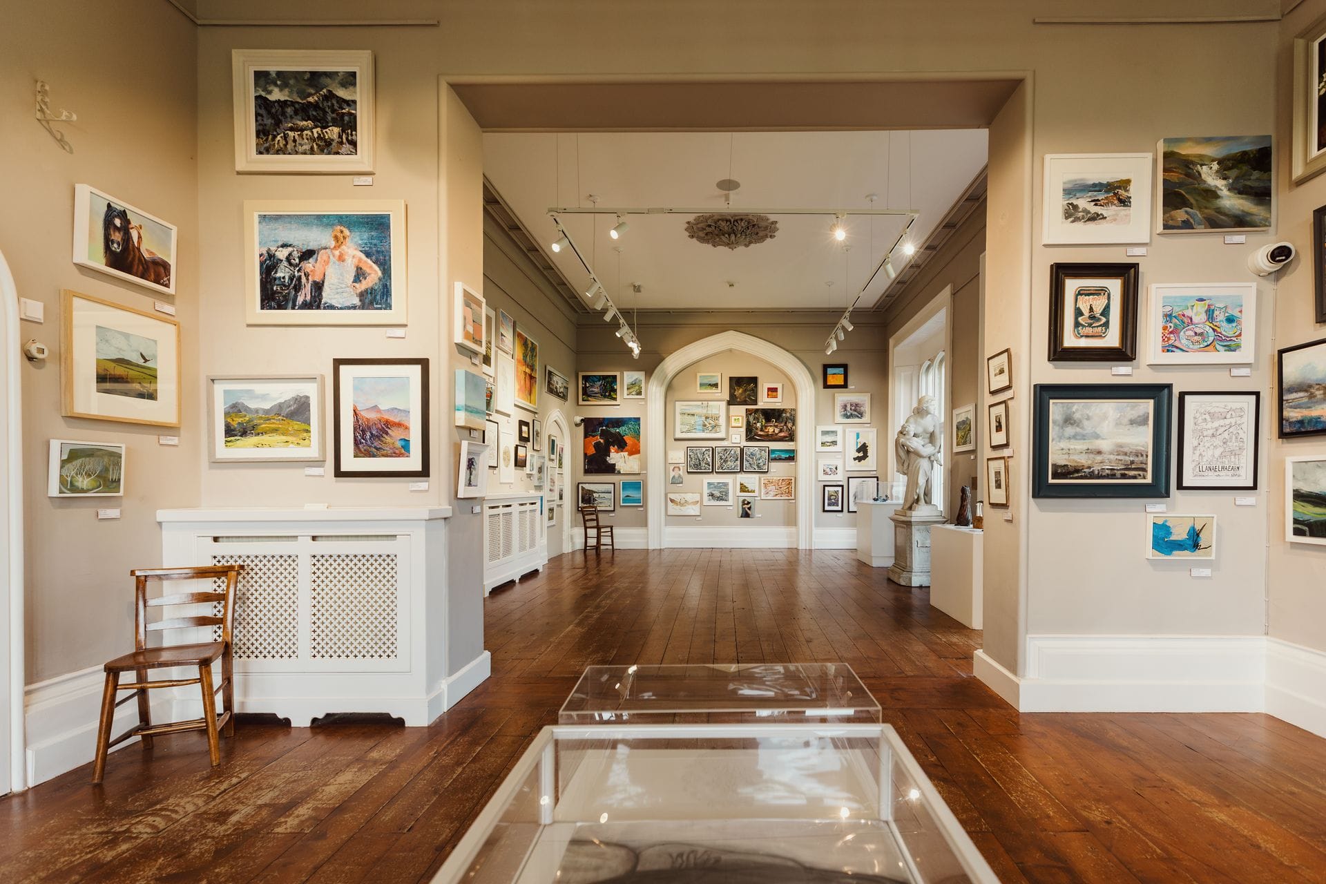 A brightly lit interior view of the Plas Glyn-y-Weddw art gallery in Wales, featuring polished wooden floors and walls beautifully covered with a diverse exhibition of framed paintings and artworks, with an archway leading to another gallery room.
