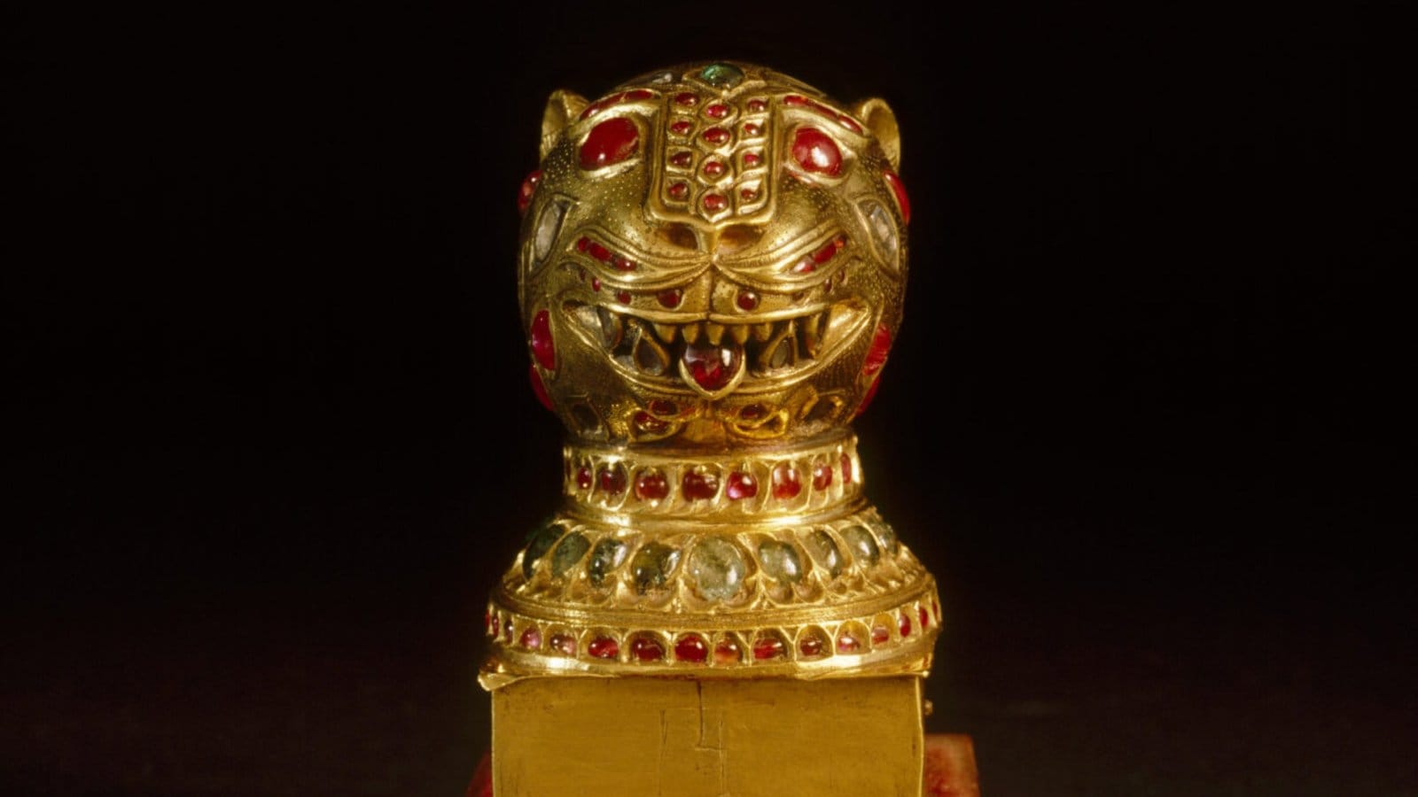 The gold tiger head finial from the throne of Tipu Sultan, set with rubies and other gems, shown close up against a black background.