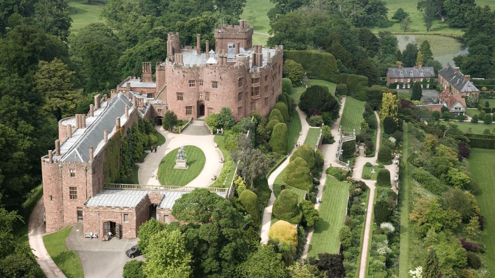 Aerial view of Powis Castle, a red sandstone castle with turrets and tall chimneys, beside formal garden terraces with clipped yew trees and lawns, surrounded by woodland and parkland.