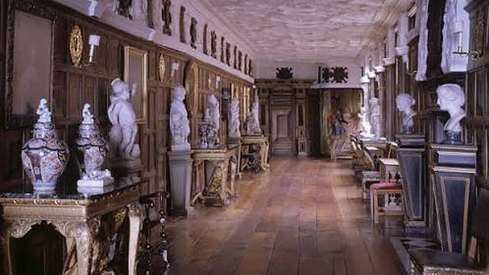 The Long Gallery at Powis Castle, a long panelled room with a polished wooden floor and plaster ceiling, lined with marble busts and statues, gilded tables and large decorative vases.
