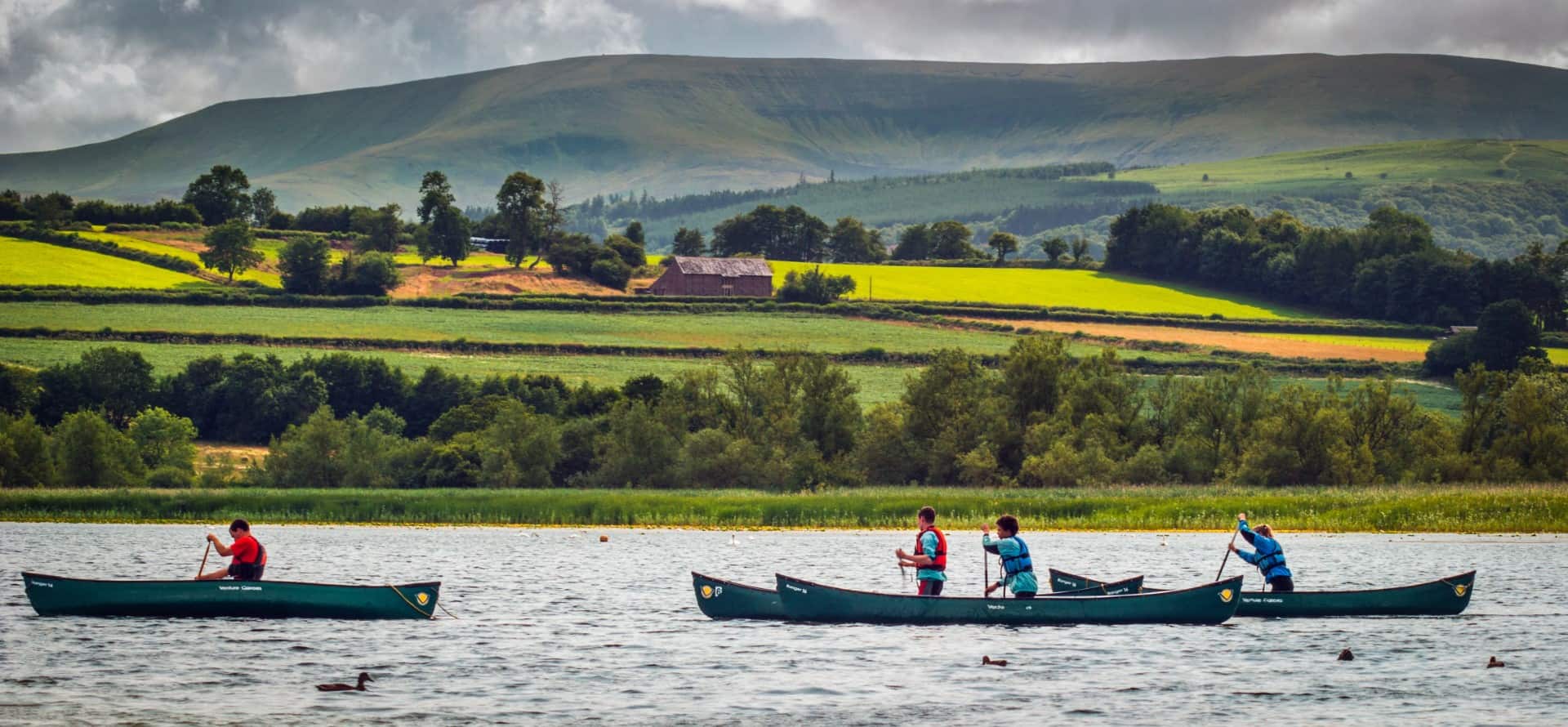 People enjoying canoeing in the Wye Valley, paddling two dark green canoes across calm water. The scenic background features a lush green shoreline, vibrant patchwork farmland on rolling hills, and a prominent mountain peak under a cloudy sky.