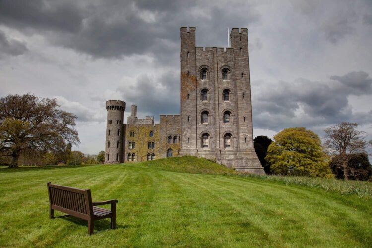 Penrhyn Castle with its tall stone towers stands on a grassy hill under a cloudy sky. A wooden bench sits in the foreground, and trees border the peaceful scene.