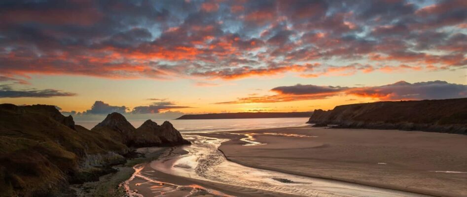 Sunset view of the wide sandy beach, rock formations, and a stream at Three Cliffs Bay on the Gower Peninsula, near Swansea, under a colorful, cloudy sky.