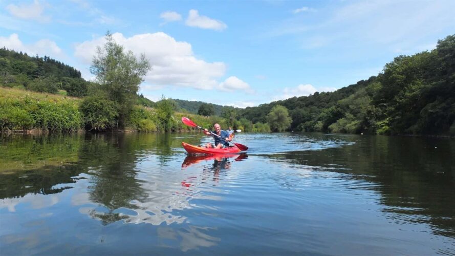 A man smiling as he paddles a red kayak, enjoying a day of canoeing on the calm, reflective waters of the River Wye. The river is bordered by lush green trees and rolling hills under a bright blue sky with fluffy white clouds.
