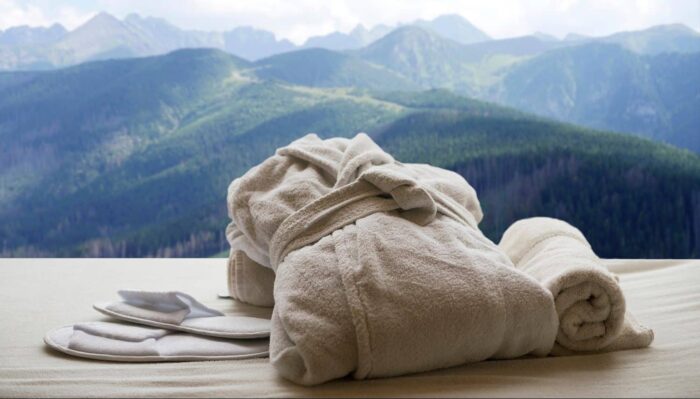 A plush white bathrobe, towel, and slippers are neatly arranged on a bed with a scenic view of green mountains and a cloudy sky—perfect for relaxing escapes in Wales.