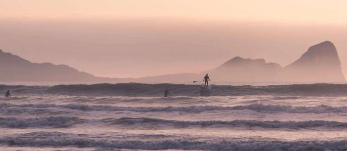 Several people surf and paddleboard on gentle sea waves at sunset, with soft pink and purple tones in the sky and distant mountains silhouetted on the horizon—a perfect evening for West Wales watersports enthusiasts.